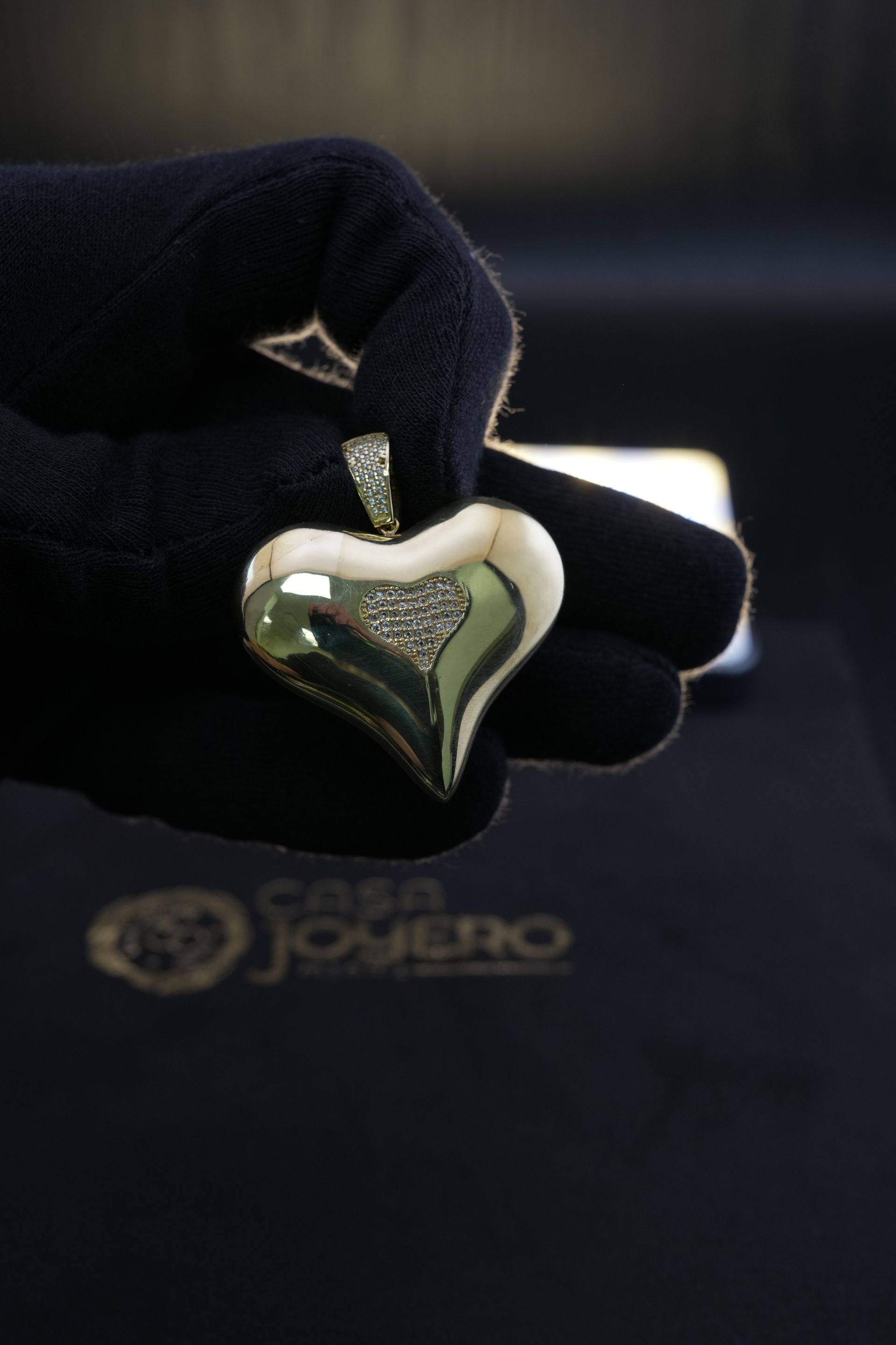 Gloved hand holding a gold heart pendant with a small diamond accent, presented on a black surface.