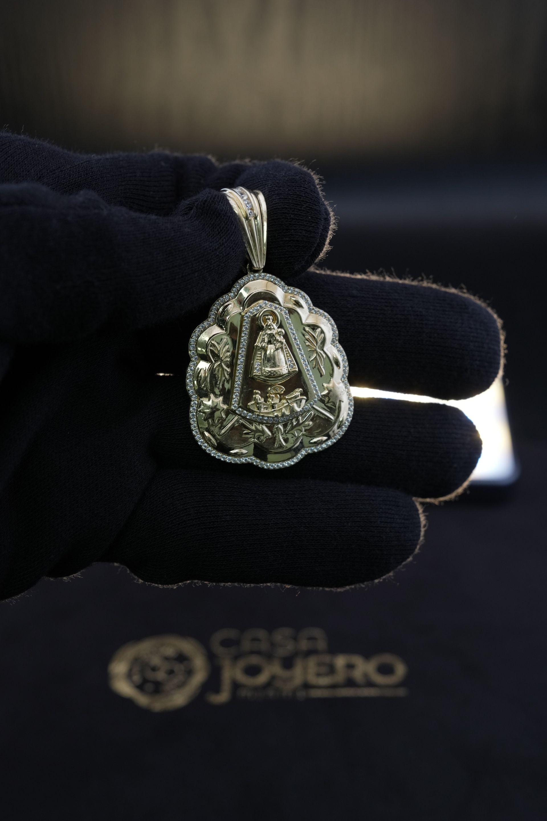Silver pendant with religious imagery held by a gloved hand, displayed on black velvet, Casa Joyero logo in the corner.