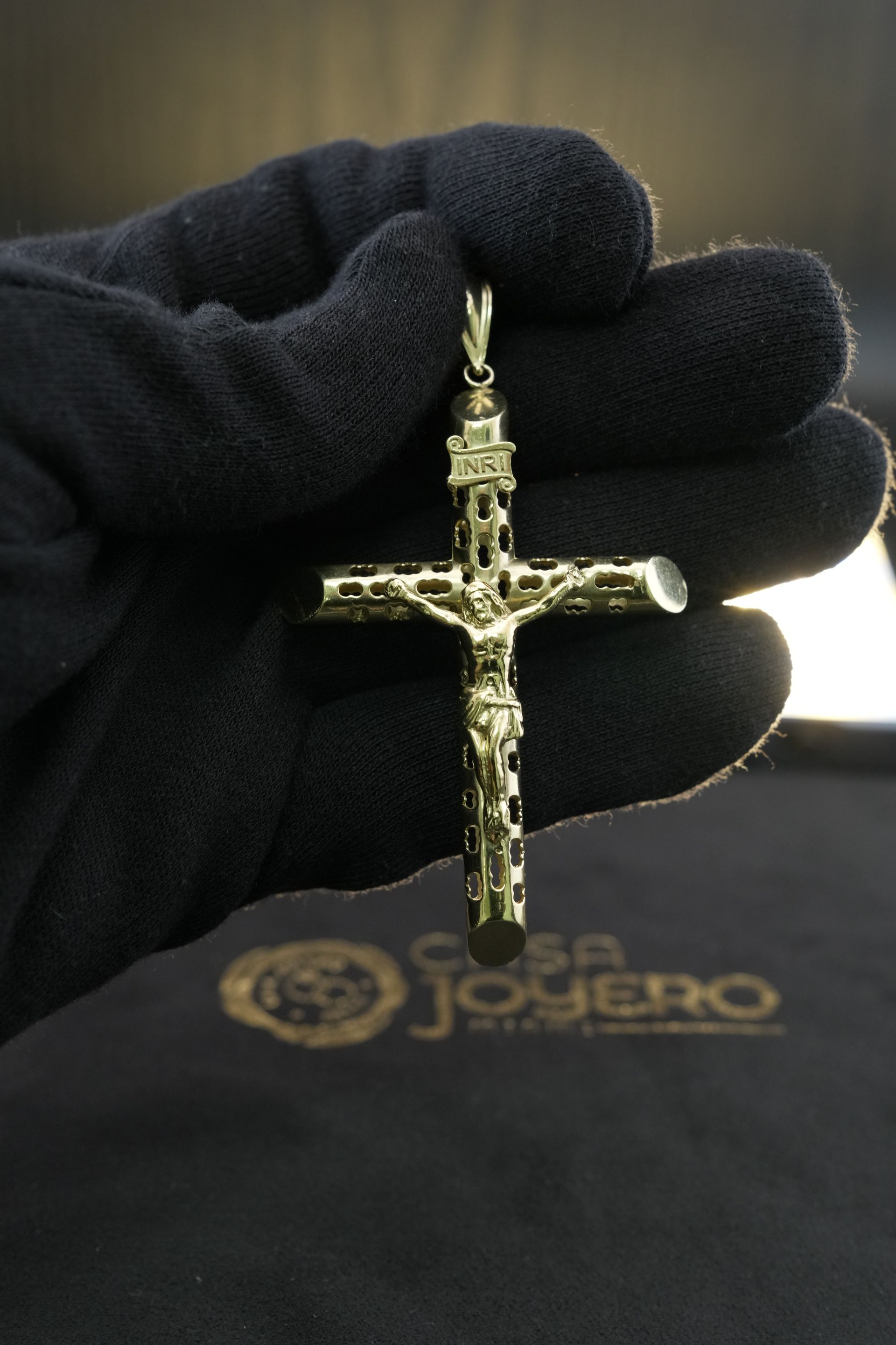 Gold crucifix pendant held in a black-gloved hand over a black cloth with a gold logo.
