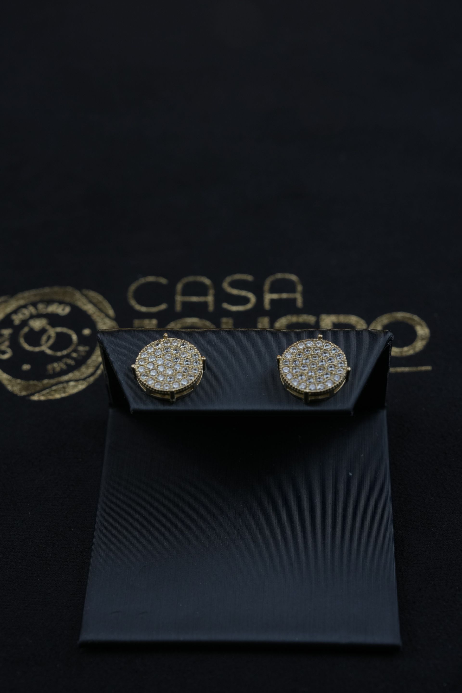 Pair of diamond stud earrings on a black display card, with a gold logo in the background.