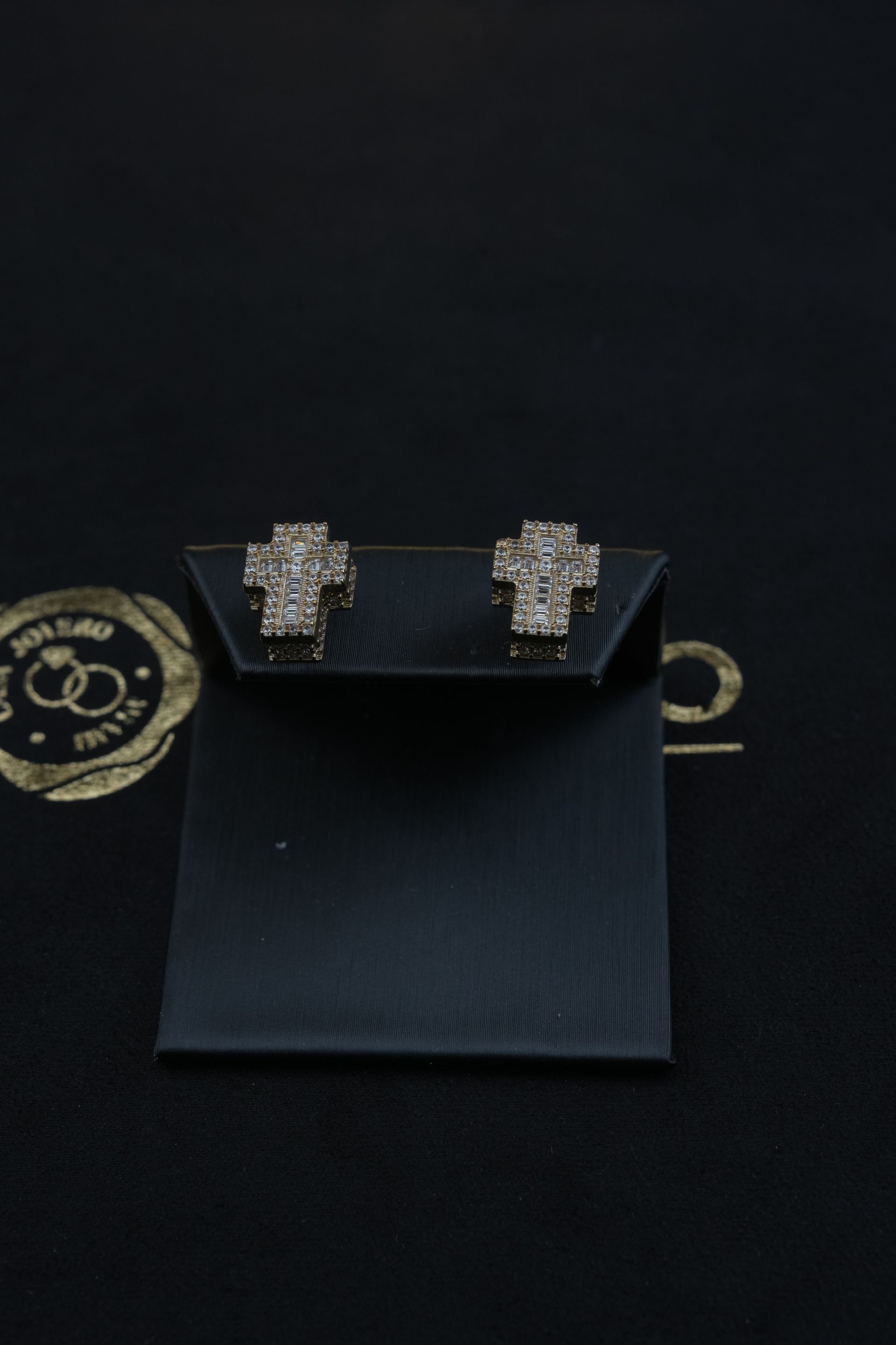 Pair of diamond cross stud earrings on a black display card, against a dark background.