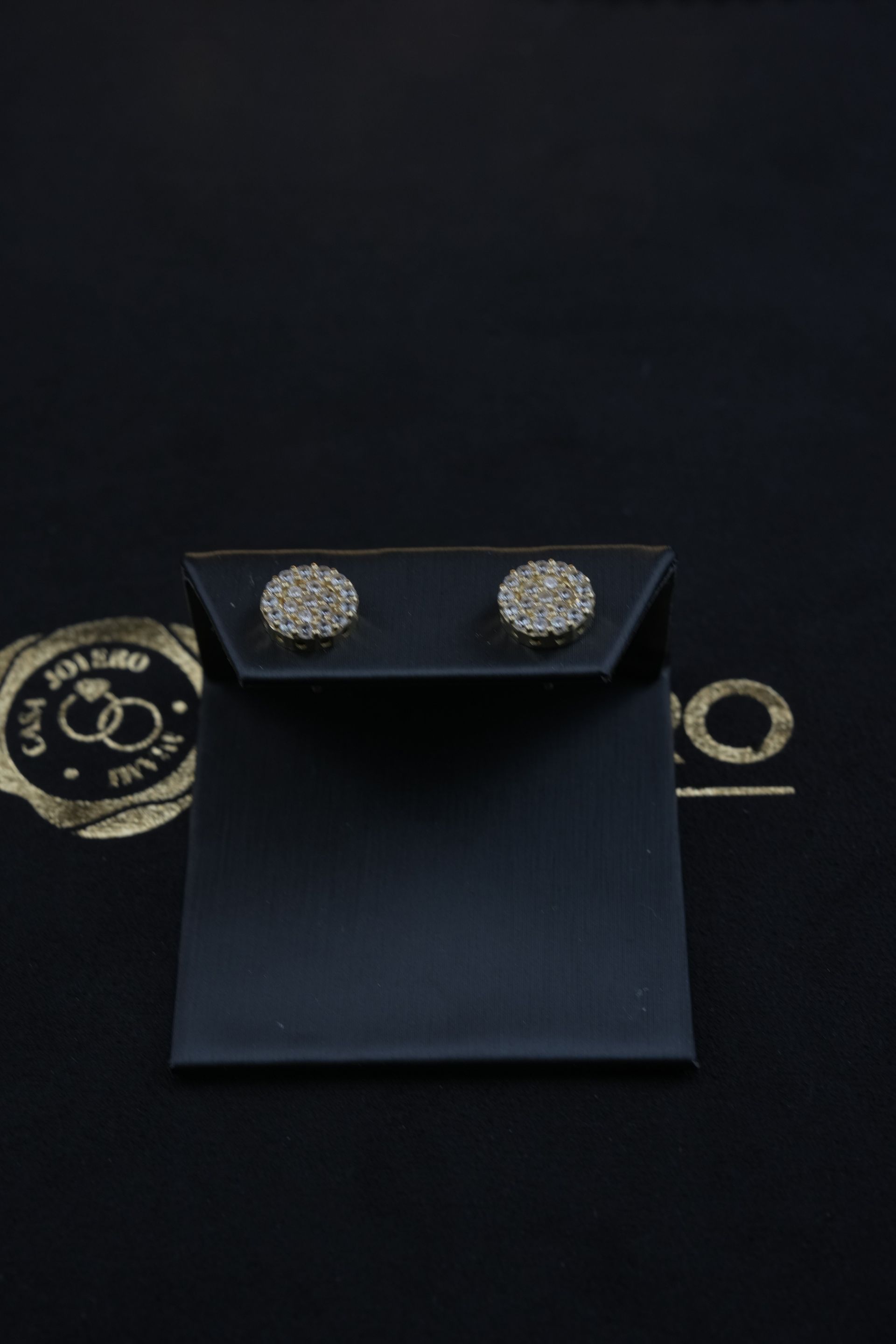 Two round, diamond-studded earrings displayed on a black earring card, set against a black background.