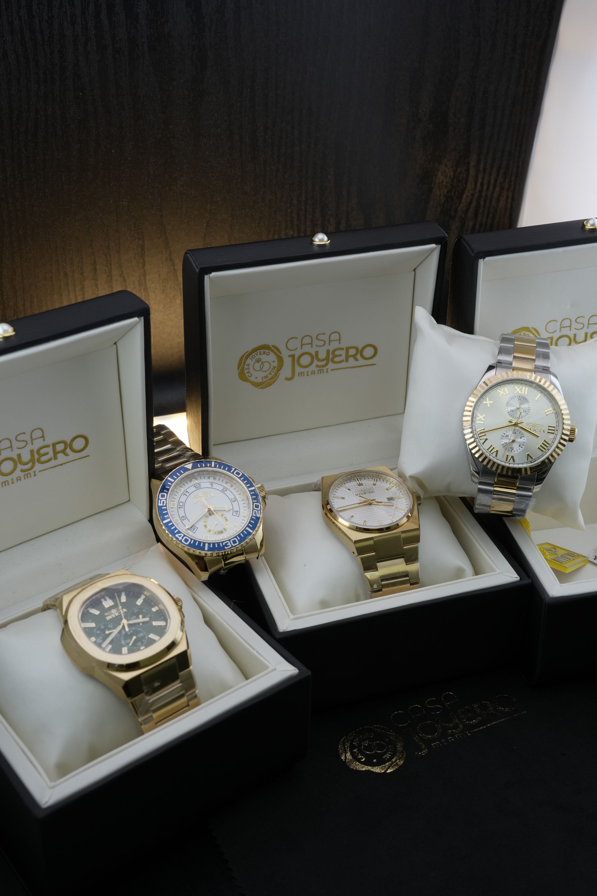 Four gold watches in open boxes, set against a dark background. Each watch sits on a white pillow with brand logos visible.