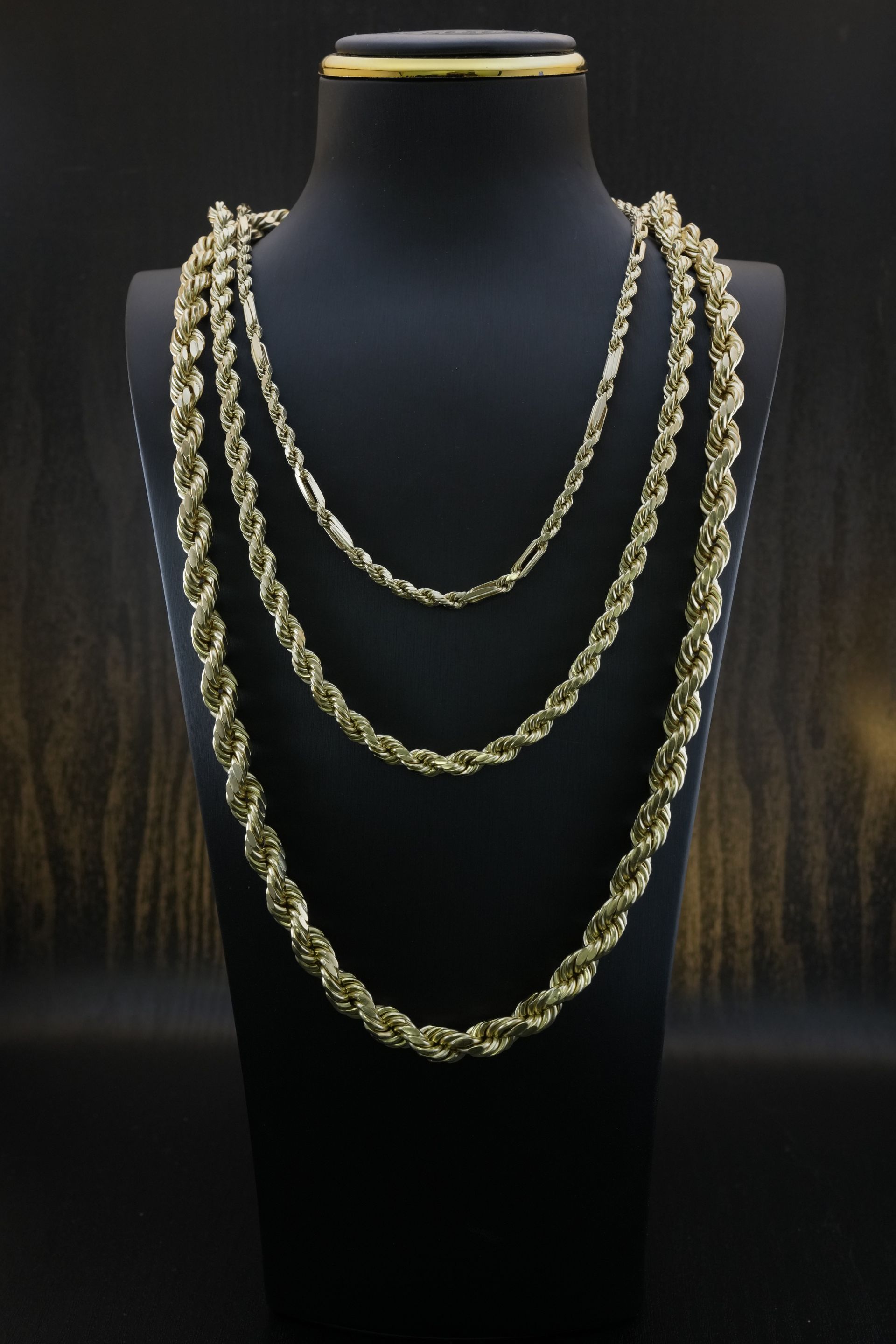 Three gold rope necklaces on a black jewelry bust against a dark backdrop.