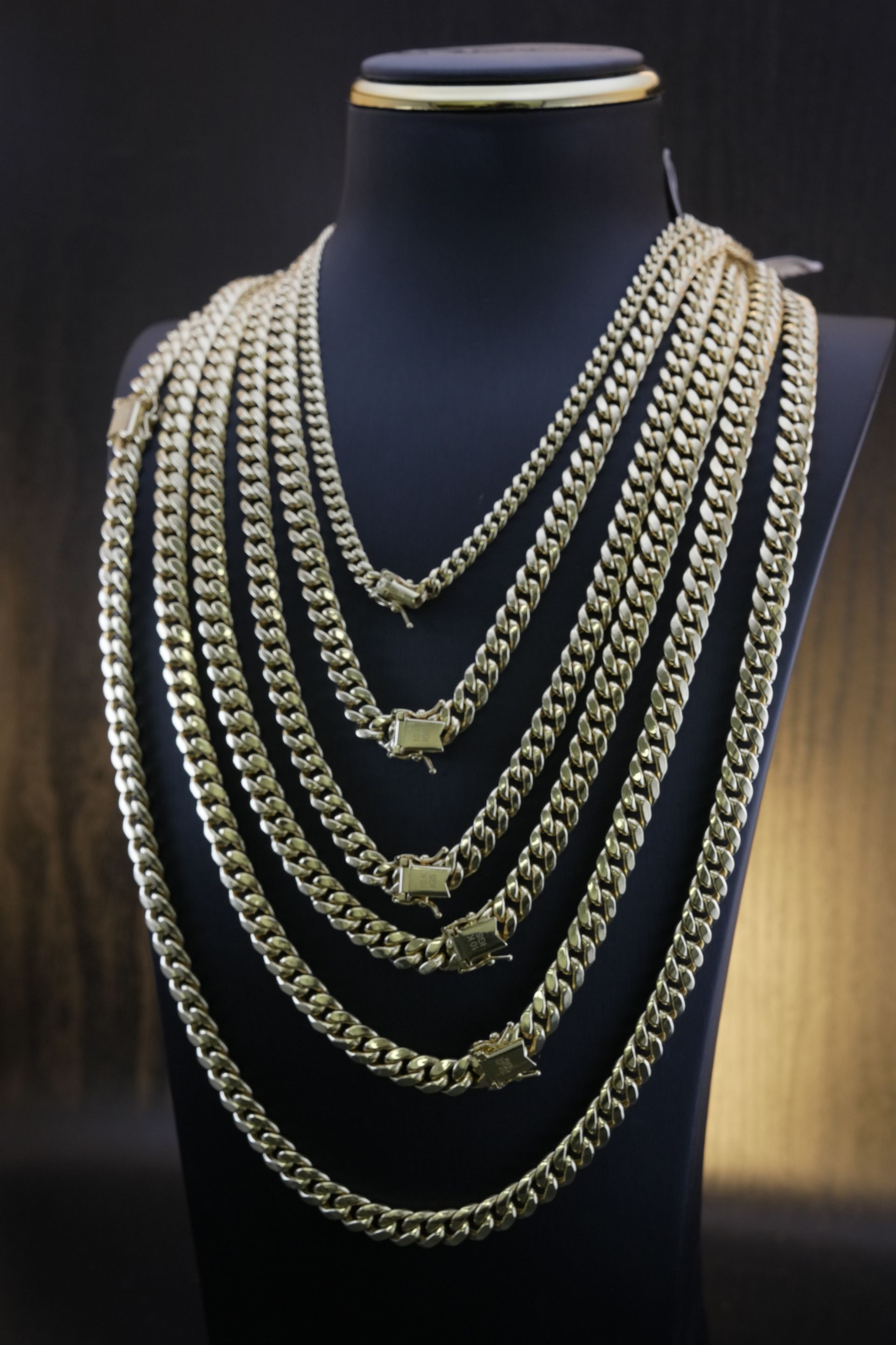 Gold curb chain necklaces displayed on a black jewelry bust in front of a dark background.