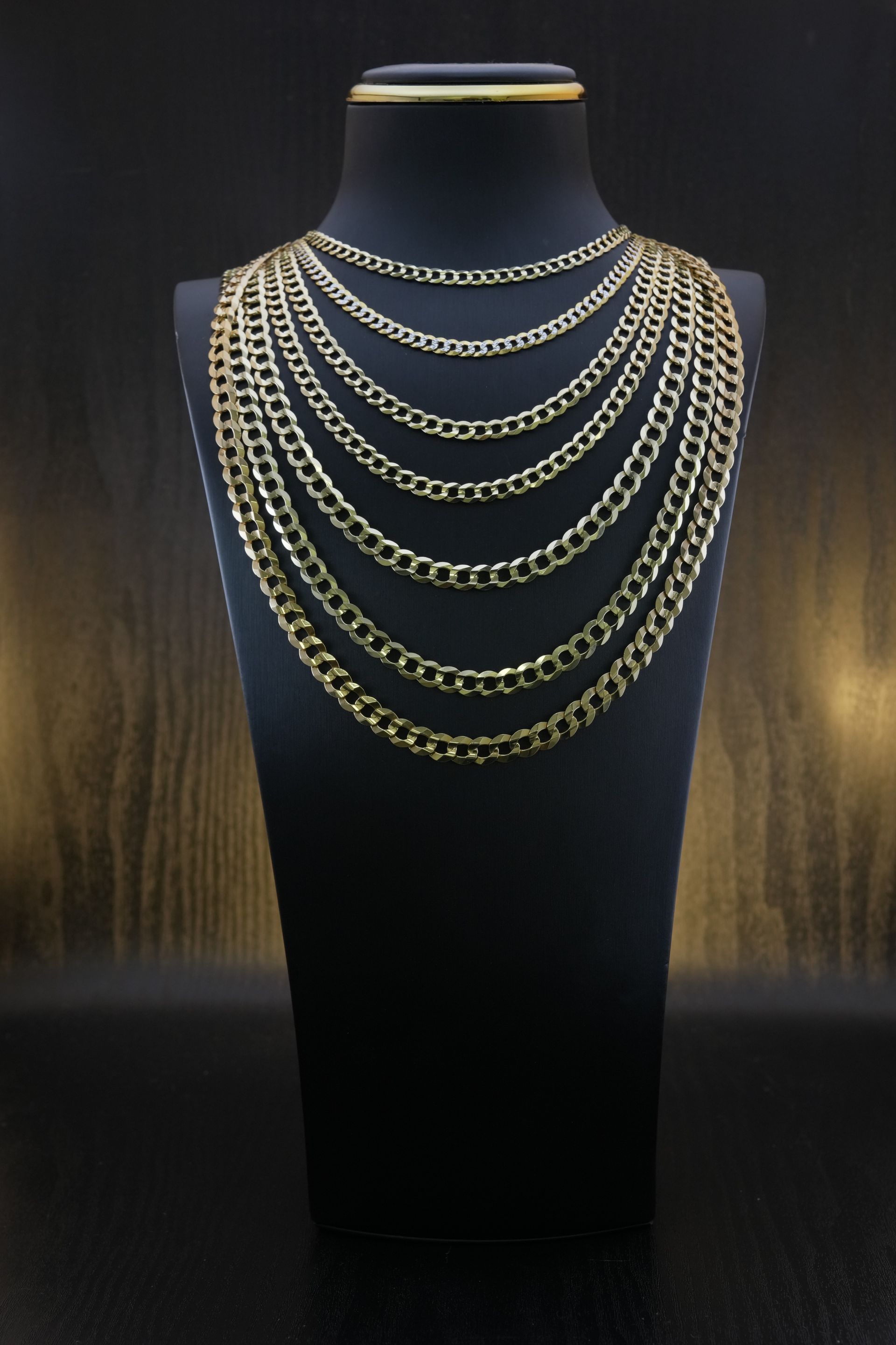 Gold chain necklaces displayed on a black mannequin against a dark background.
