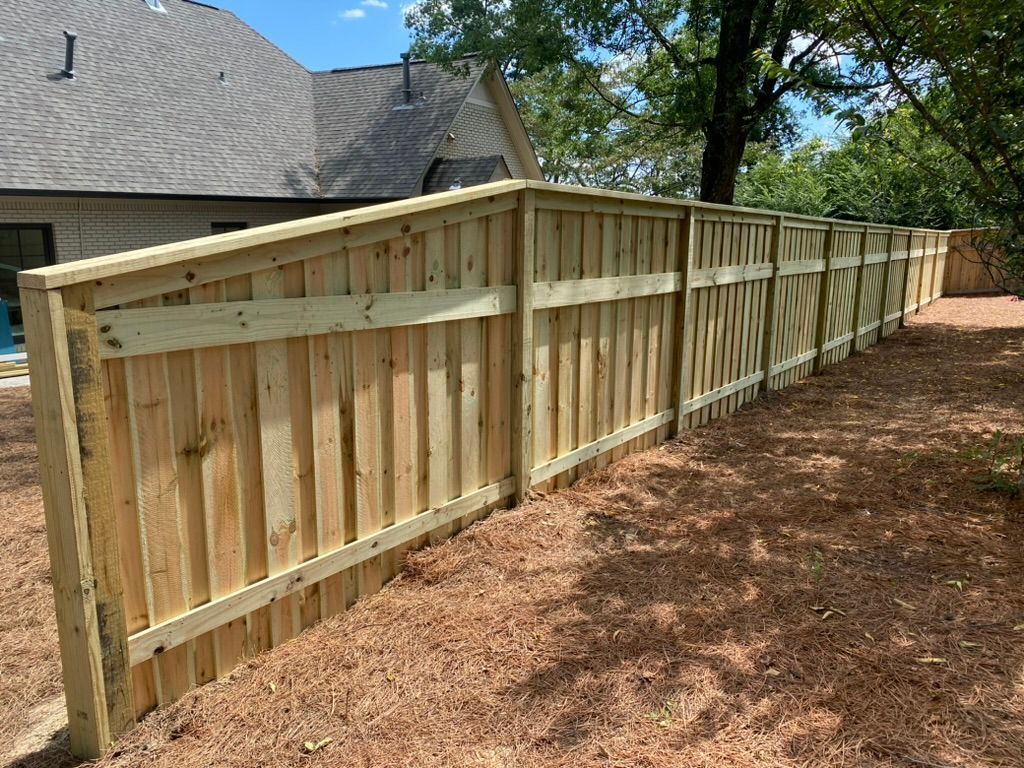 A newly constructed, light-colored wooden fence standing along a yard covered in pine straw with a house in the background.