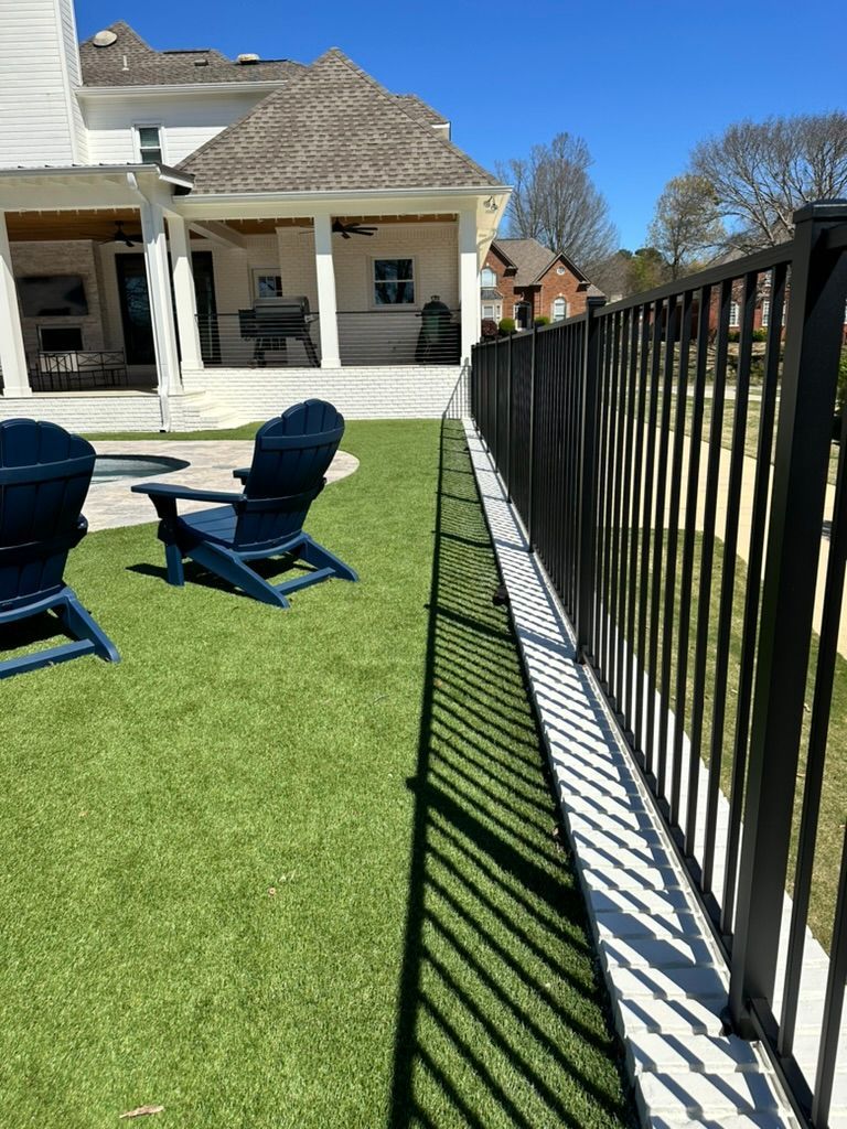 Two blue Adirondack chairs on a grassy lawn next to a metal fence casting a long shadow toward a house patio.