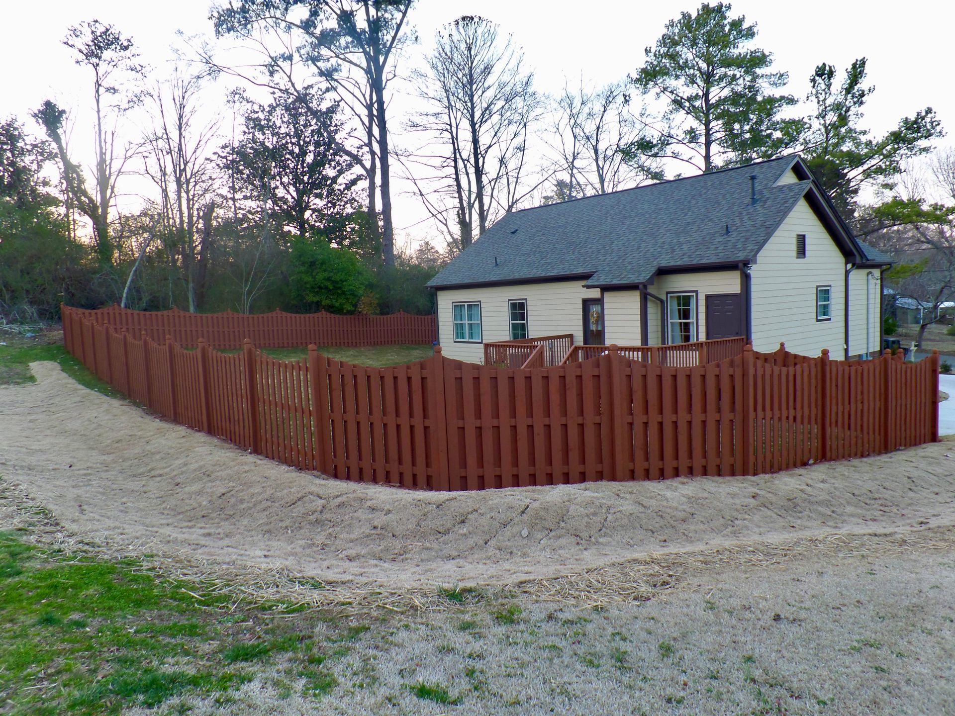 A light-colored house with a dark roof stands behind a decorative, reddish-brown scalloped wooden picket fence.