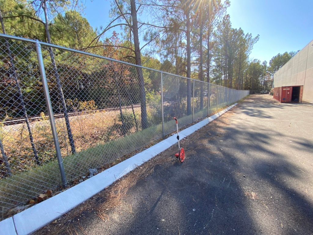 A chain-link fence borders a paved parking lot with a freshly installed white concrete curb and a red measuring wheel.