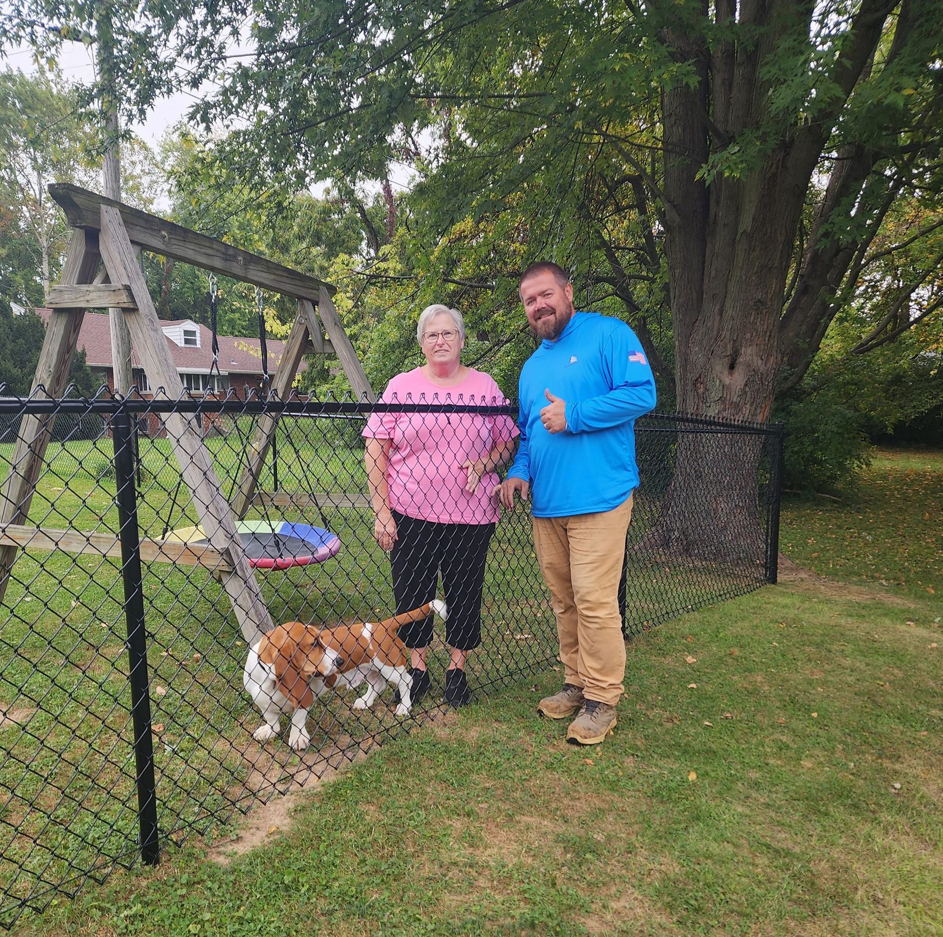 A person in a pink shirt and another in a blue shirt stand behind a chain-link fence with a Basset Hound on the grass.