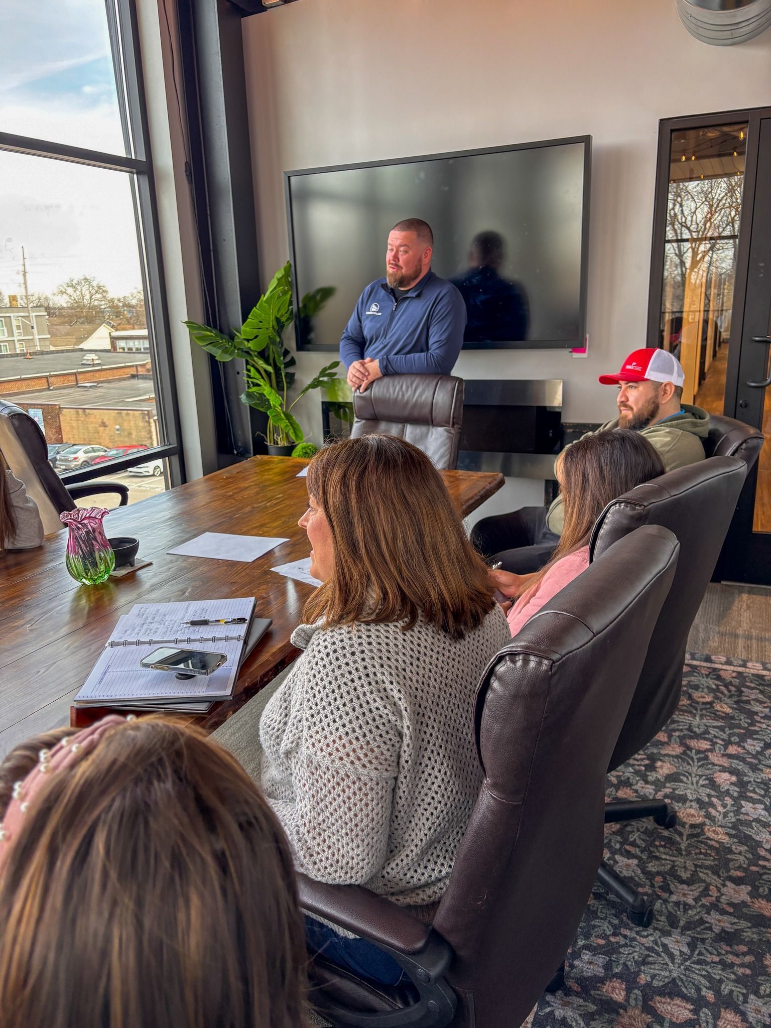 A man stands at the head of a conference table leading a meeting with four people seated in a sunlit office space.
