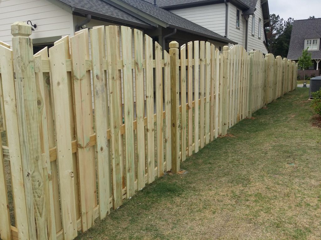 A new, light-colored wooden picket fence with a scalloped top design follows the edge of a residential lawn.