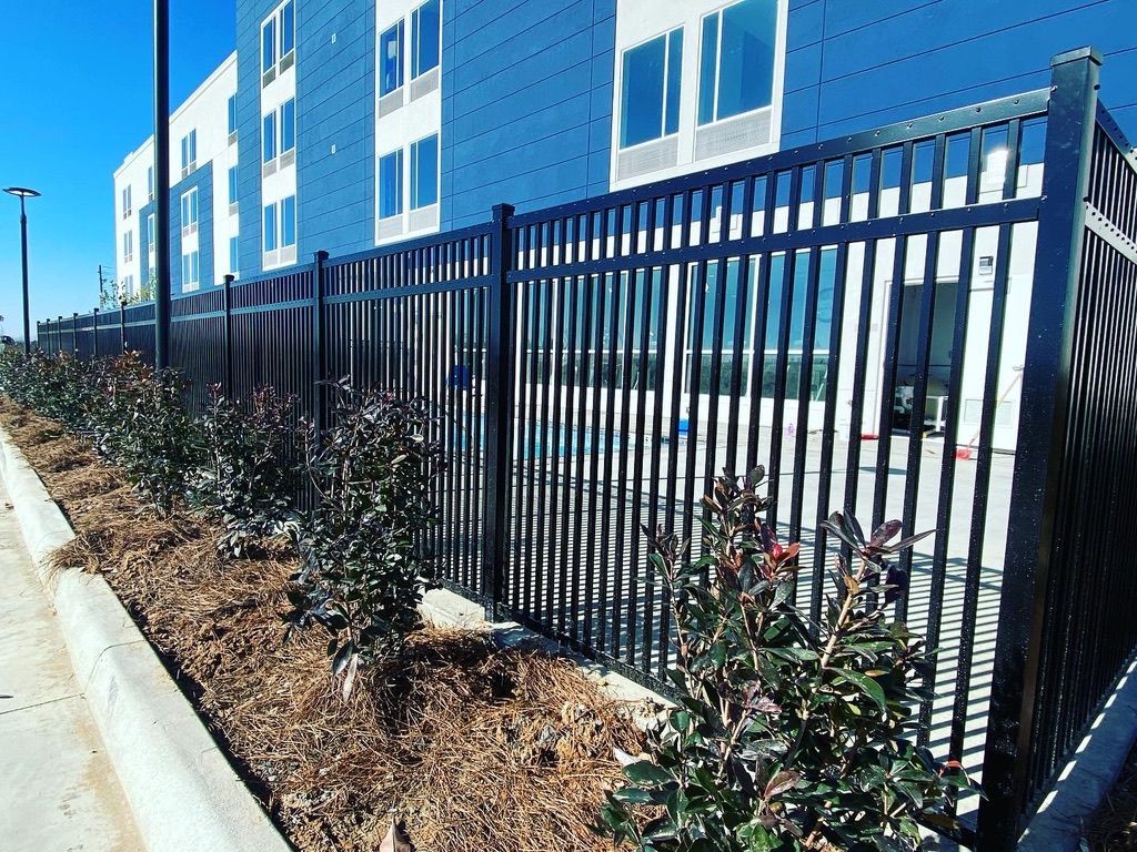 A tall black metal security fence lines a sidewalk with a mulch garden bed and a blue and white building in the background.