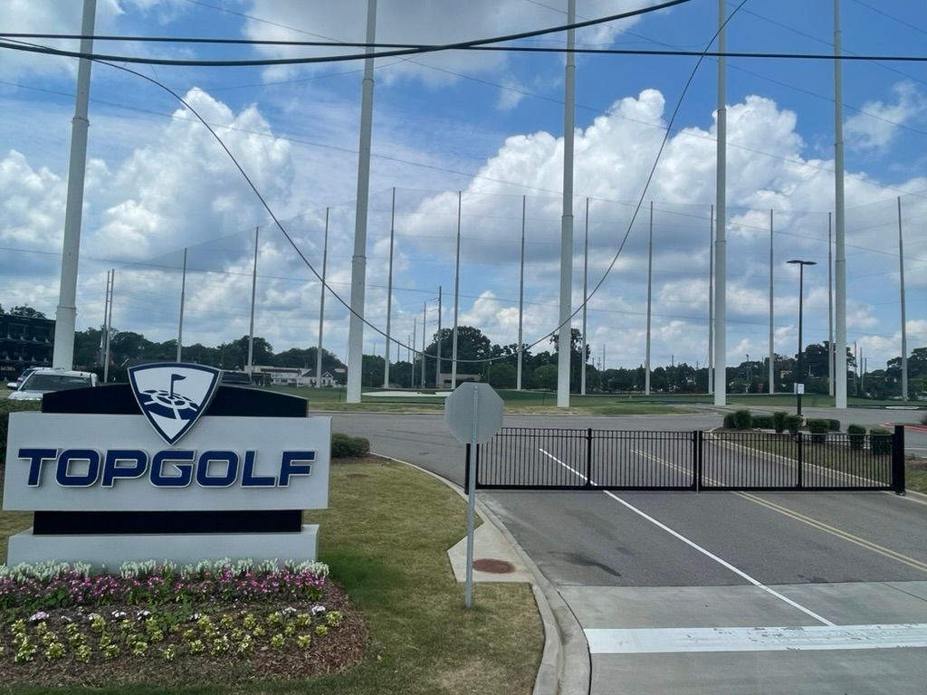 The Topgolf entrance sign in white and blue, with tall safety netting poles visible against a blue, cloudy sky.