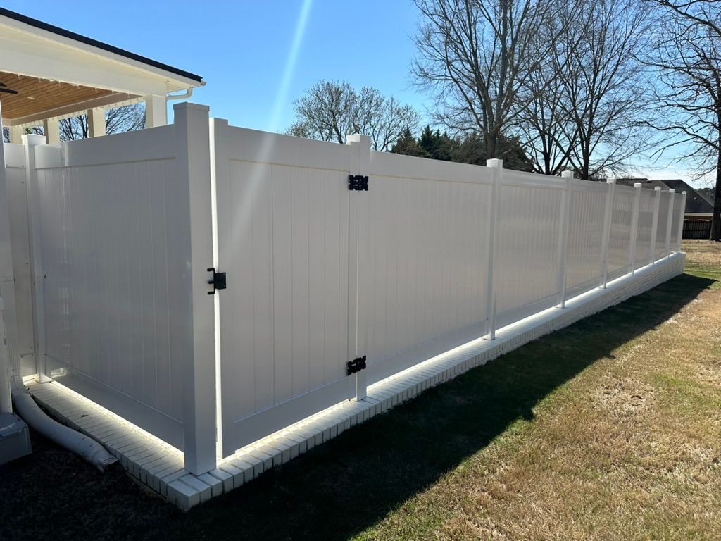 A white vinyl privacy fence with a gate, installed on a low brick base in a grassy yard under a clear blue sky.