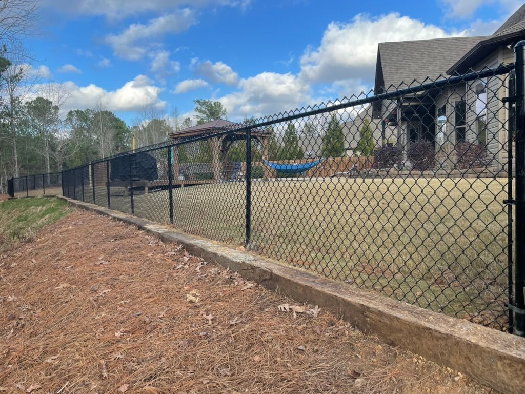 A black chain-link fence mounted on a low concrete retaining wall borders a residential lawn with a house and gazebo.