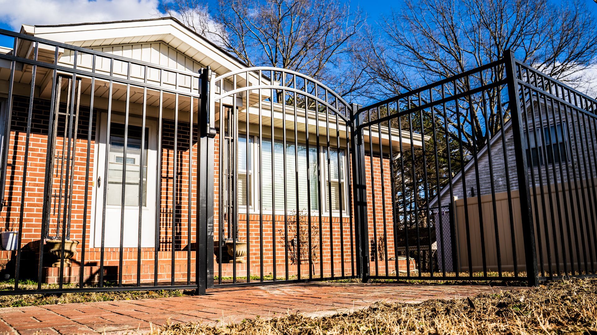 A black metal arched gate stands in front of a brick house with a white door and window under a bright blue sky.