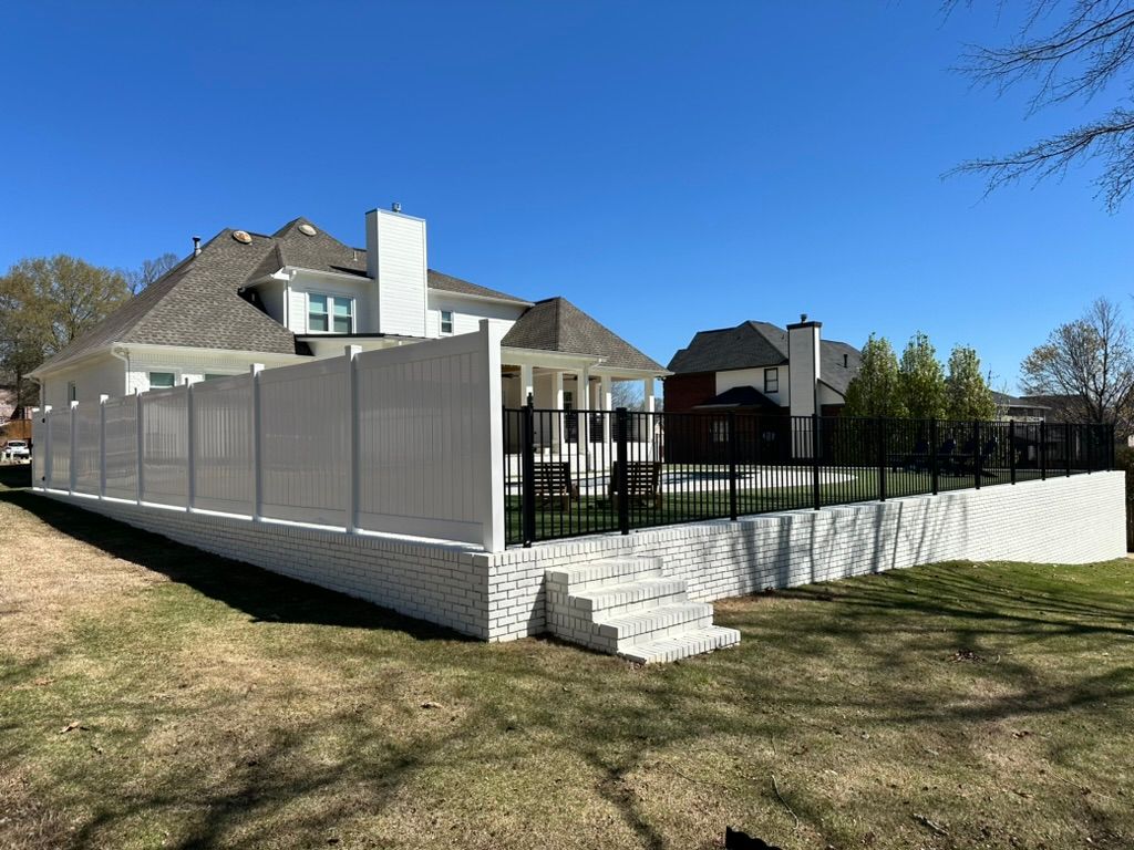 A white brick retaining wall with steps surrounds a backyard featuring a white privacy fence and a black metal fence.