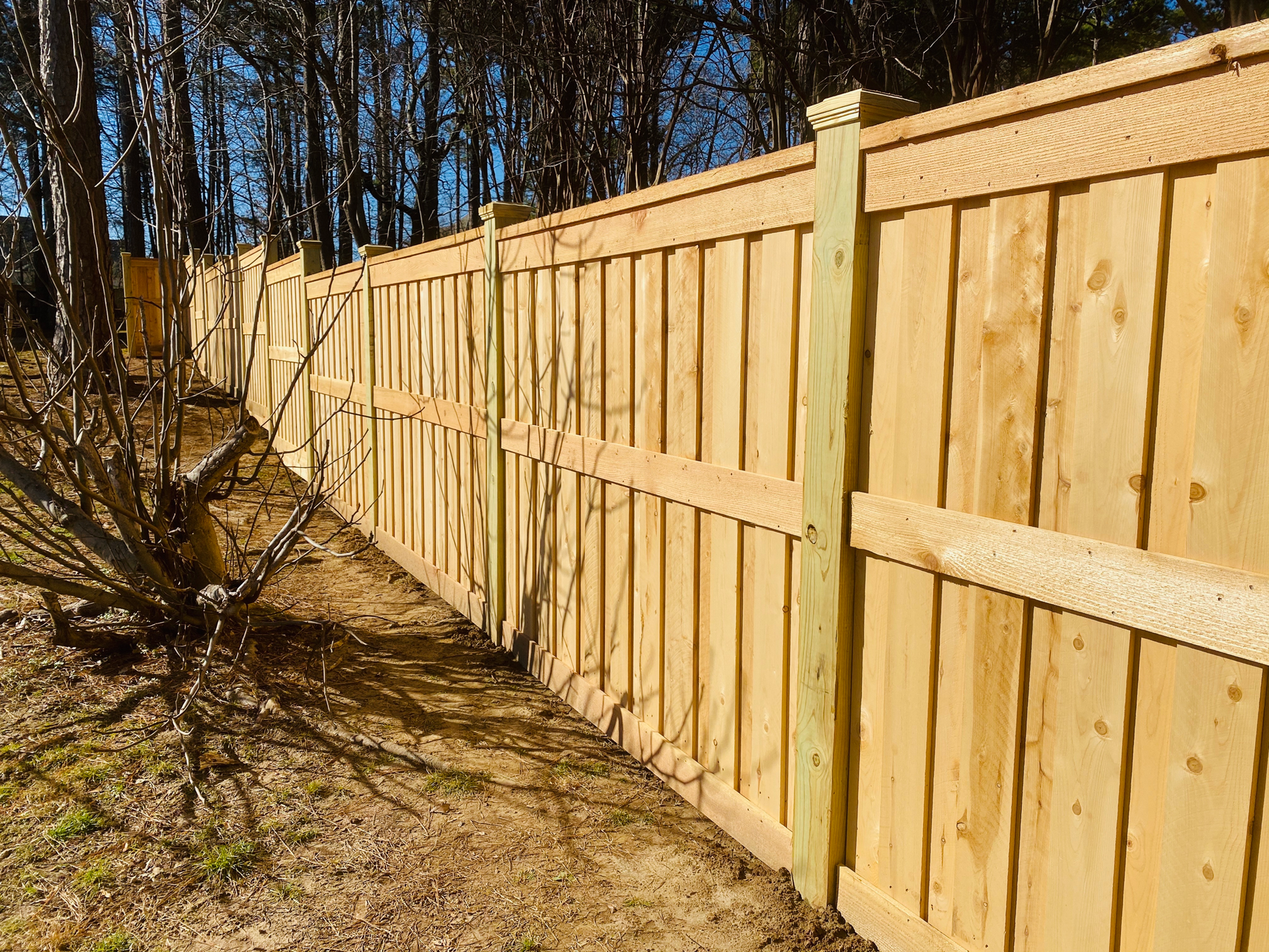 A tall, light-colored wooden privacy fence stretches along a dirt ground path with trees in the background.