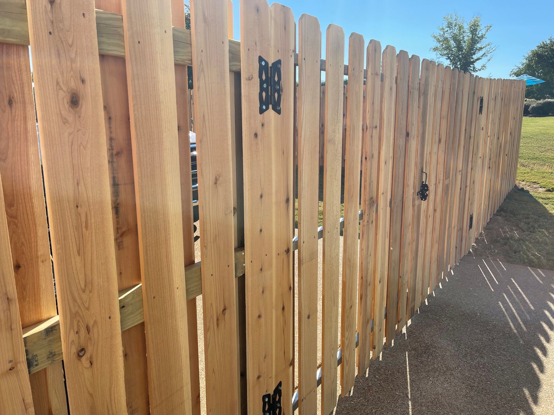 A perspective view of a new wooden fence with dark metal gate hinges, stretching toward the background on a sunny day.