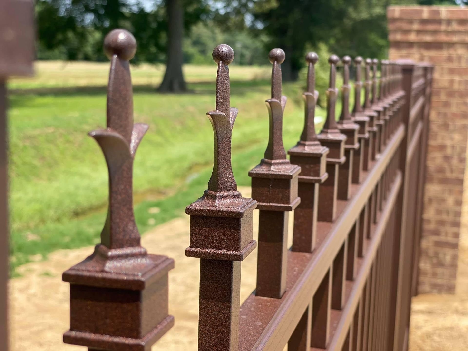 A close-up of a bronze-colored metal fence with ornate, spear-like finials attached to square posts in an outdoor setting.