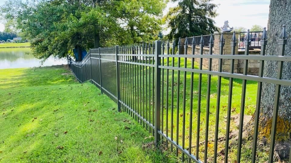 A black metal picket fence runs through a grassy yard alongside a lake, leading toward a stone patio under trees.