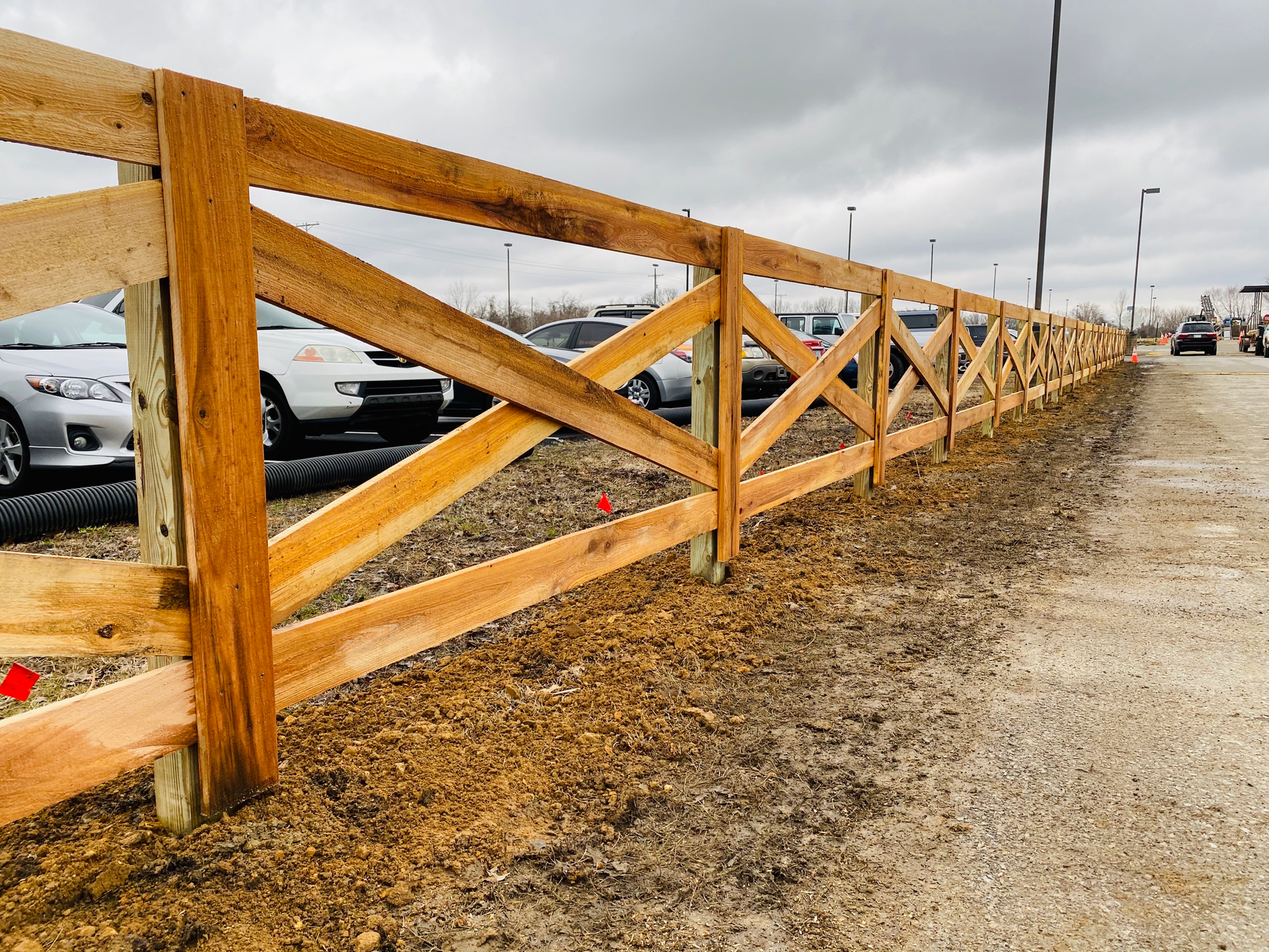 A wooden cross-buck fence runs alongside a parking lot with several parked cars under a cloudy sky.