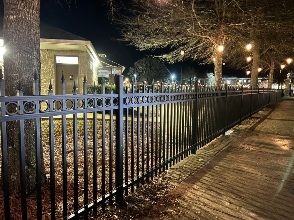 A black metal ornamental fence runs along a paved path next to a building and trees at night.