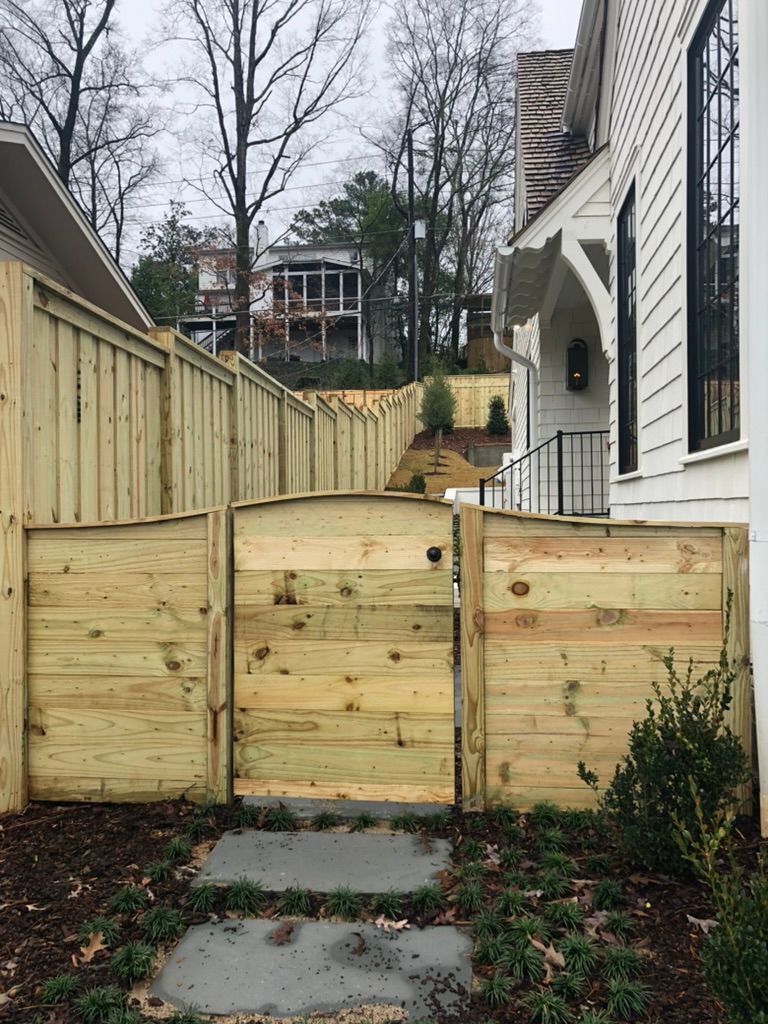 A wooden privacy fence with a matching arched gate, situated on a stone pathway next to a white house.