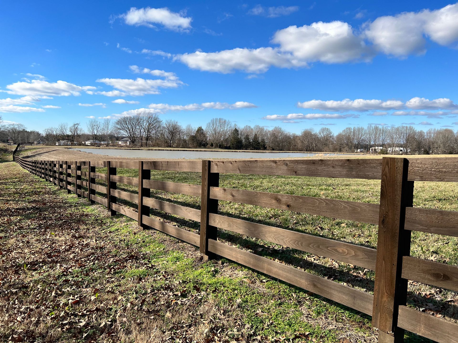A long, dark wooden fence runs through a grassy field under a bright blue sky with scattered clouds.