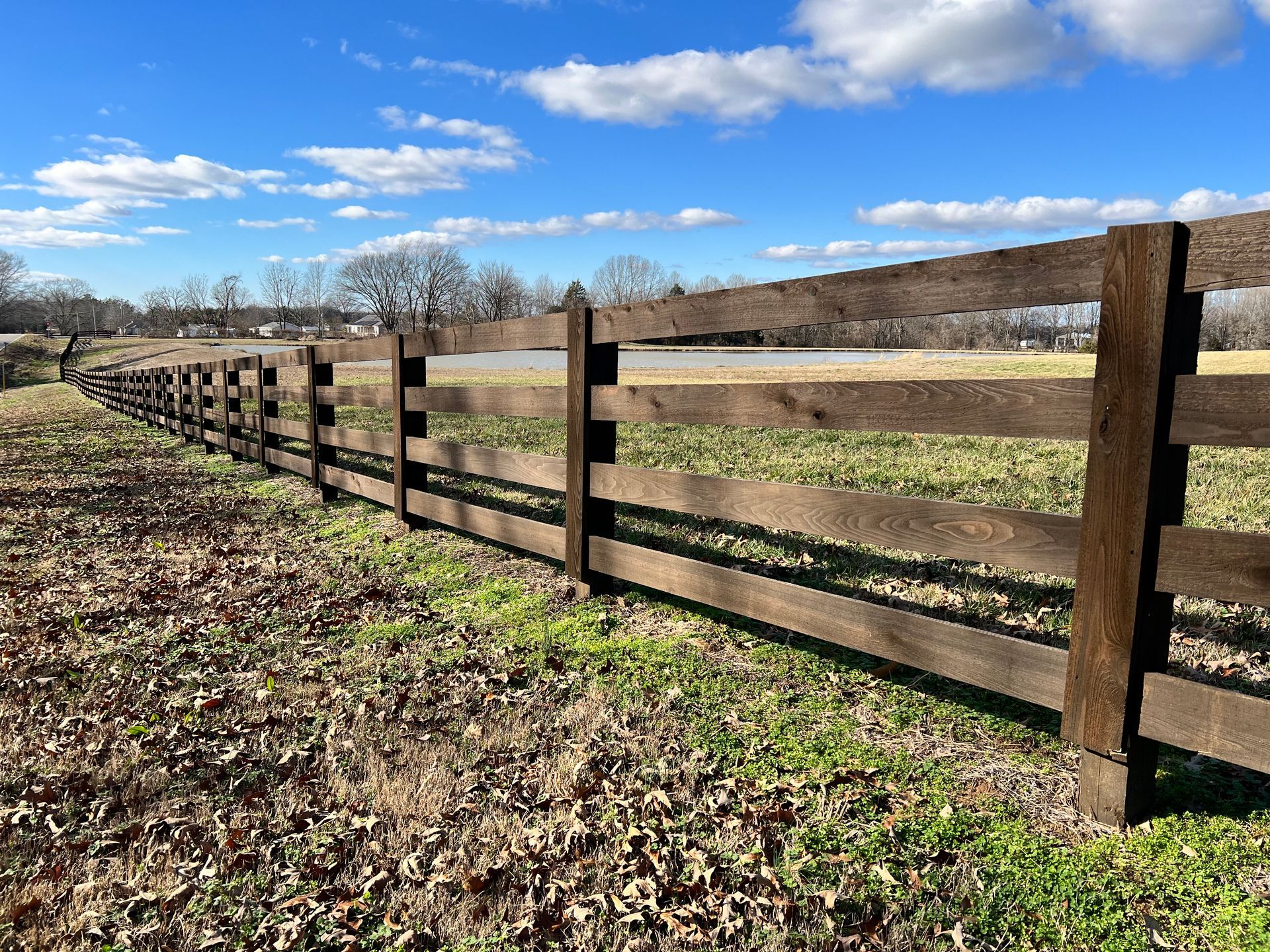 A dark wooden rail fence runs diagonally across a rural field under a bright blue sky with scattered clouds.