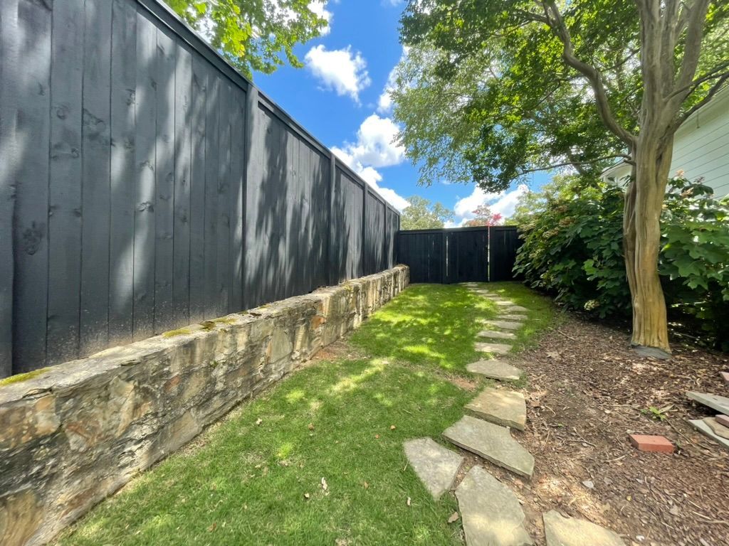 A stone pathway runs through a grassy yard alongside a tall, dark wooden fence and a stone retaining wall.