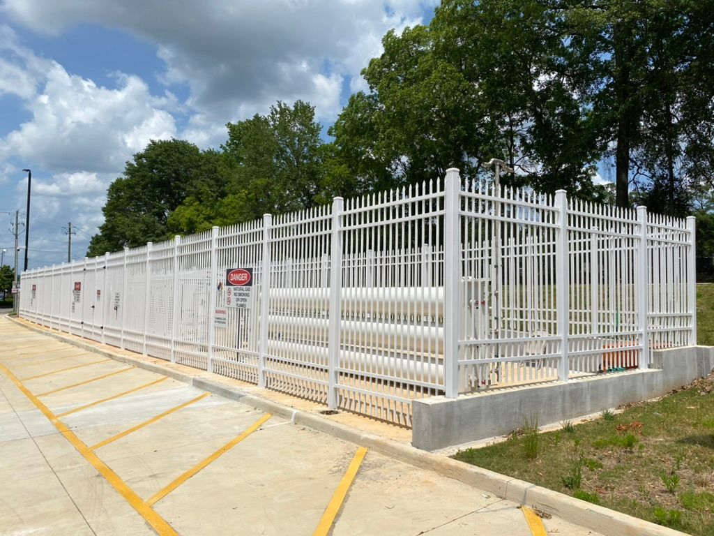 A fenced white utility installation with large horizontal pipes behind it, located next to a parking lot under a blue sky.