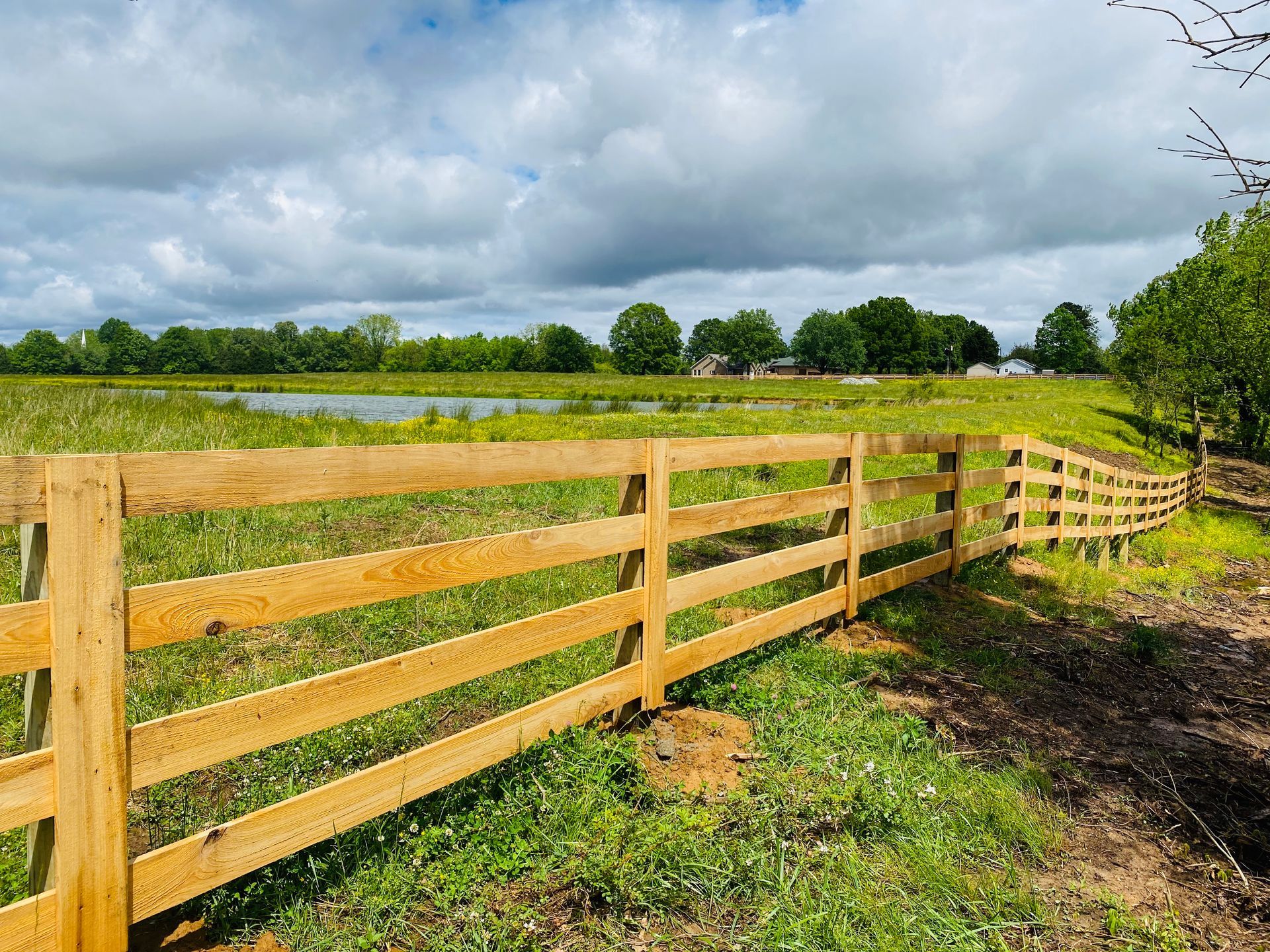 A wooden post-and-rail fence stretches across a green pasture toward a distant pond and tree line under a cloudy sky.