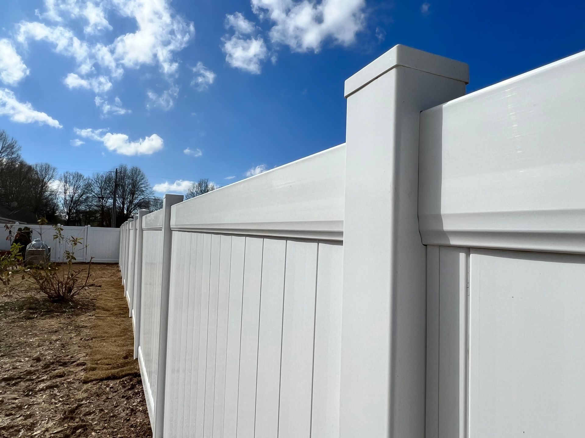 A white vinyl privacy fence stands outdoors against a bright blue sky with scattered white clouds.