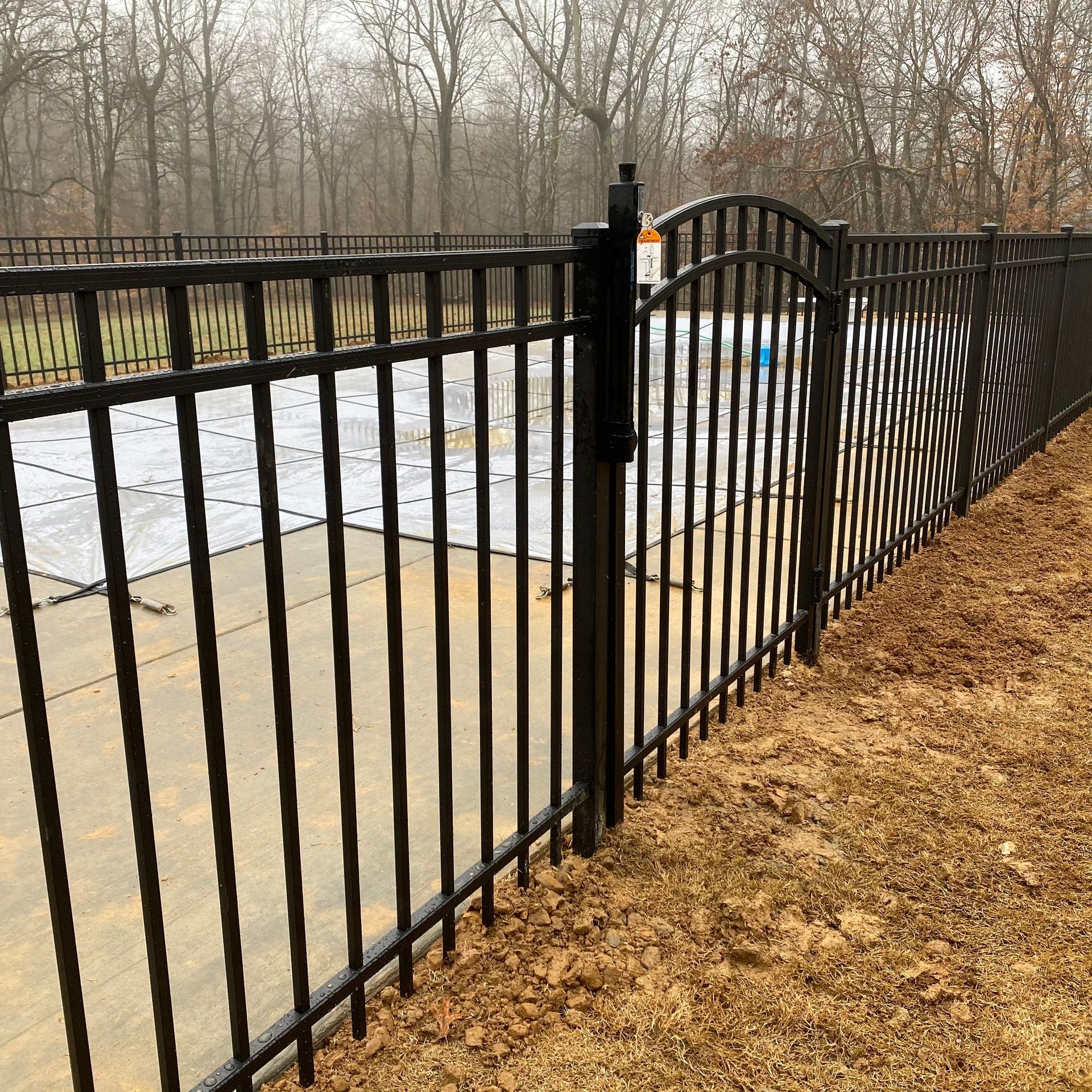 A black metal pool fence and arched gate stand on a concrete pool deck with a covered swimming pool in the background.