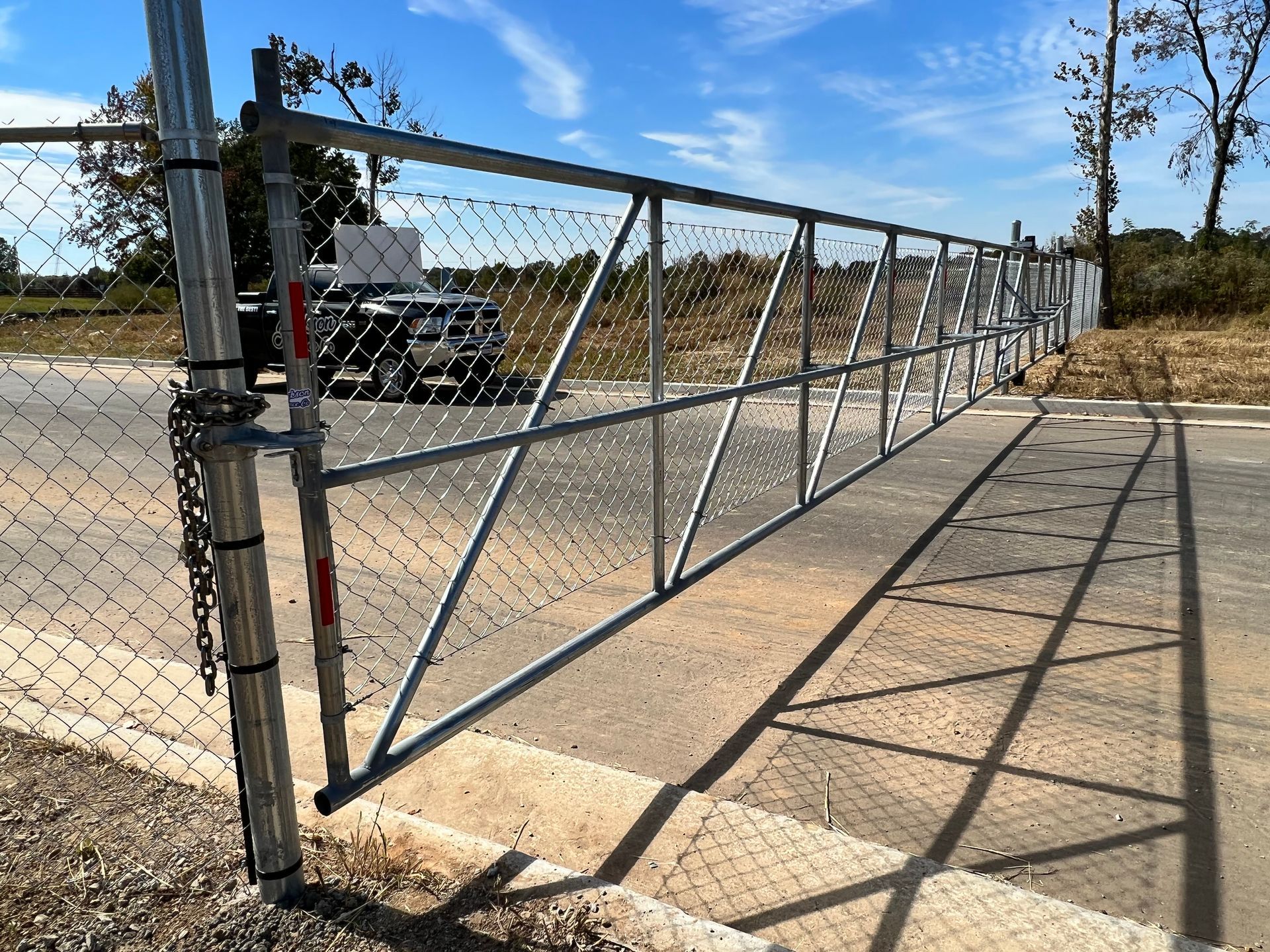 A metal chain-link gate secured with a chain stands across a paved road under a bright blue sky.