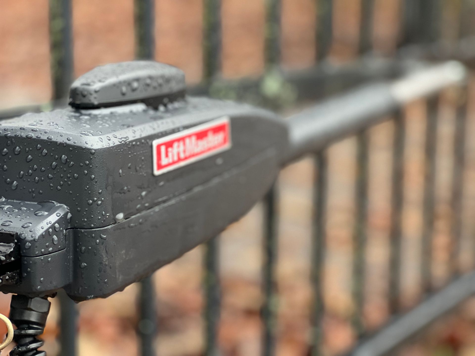 A wet, dark gray LiftMaster gate opener arm mounted to a black metal fence.