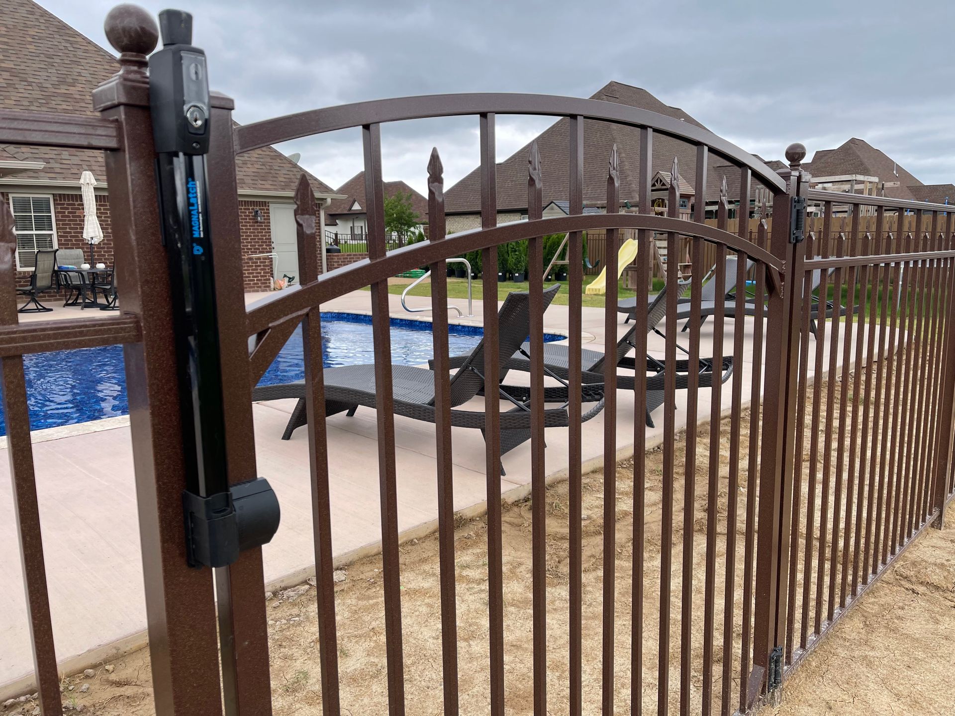 A brown metal fence with a gate equipped with a black automatic latch mechanism, overlooking a backyard swimming pool.