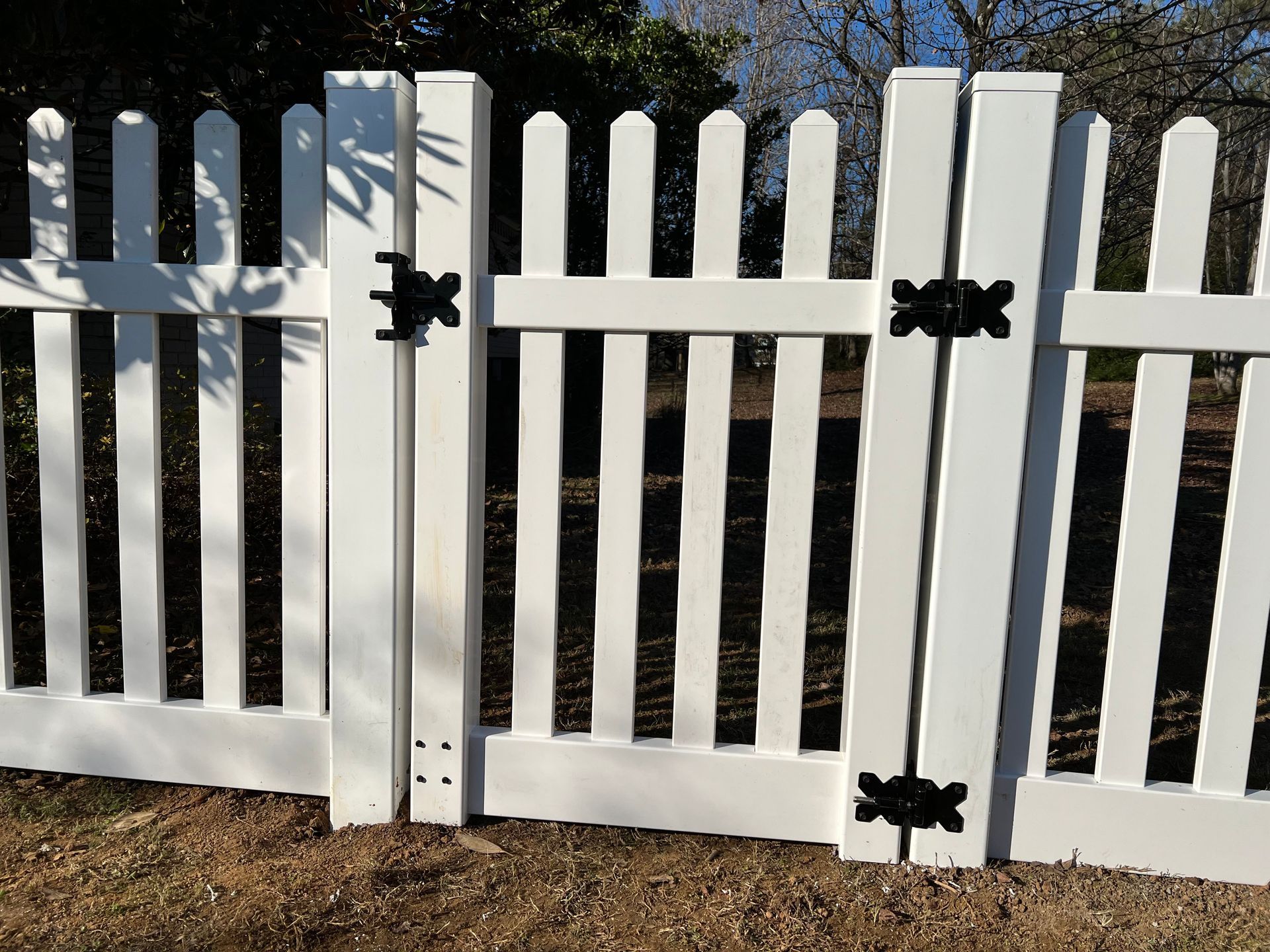 A white vinyl picket fence with a double gate featuring black decorative hardware, set against a blurred outdoor background.