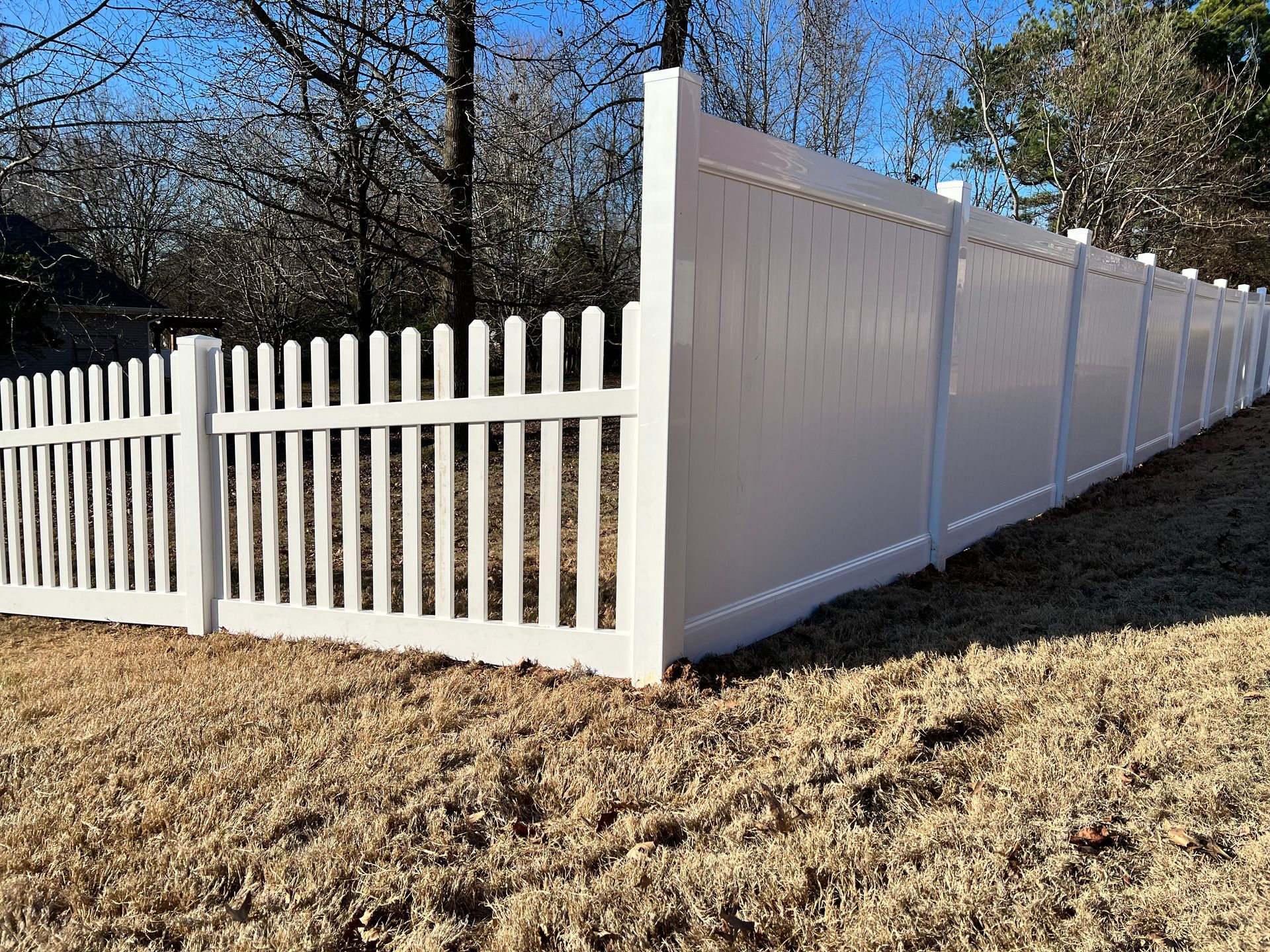 White vinyl fence transitioning from a short picket style to a tall, solid privacy wall on a grassy slope.