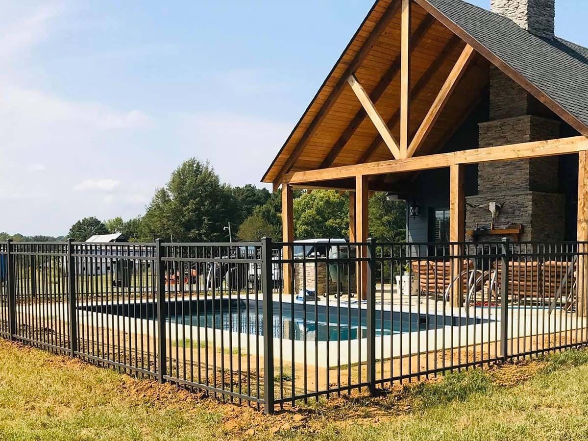 An outdoor backyard pool enclosed by a black metal fence next to a wooden open-air patio structure with a stone fireplace.