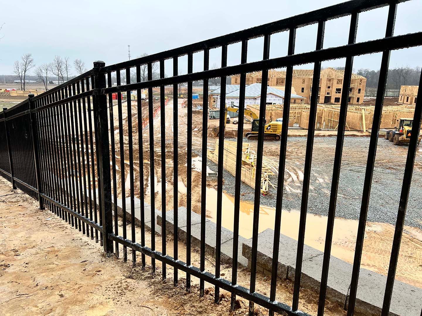 Black metal fence overlooking an active construction site with buildings, dirt, and heavy machinery under an overcast sky.