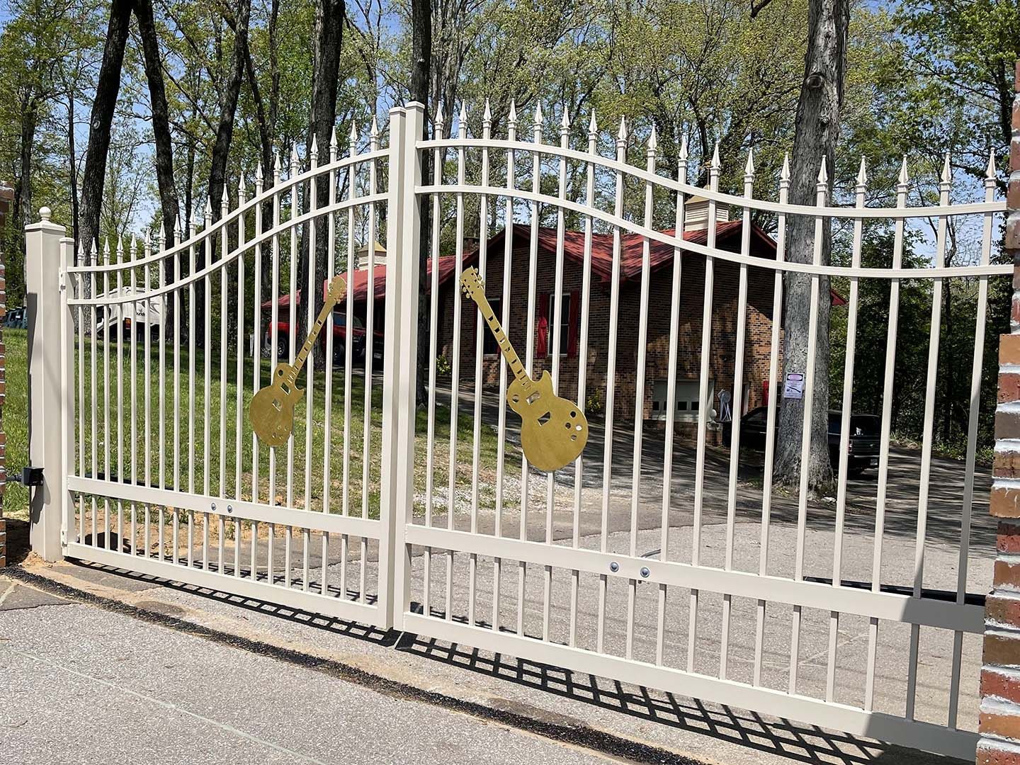 Off-white metal gate with decorative gold electric guitar cutouts, set against a wooded background with a house.