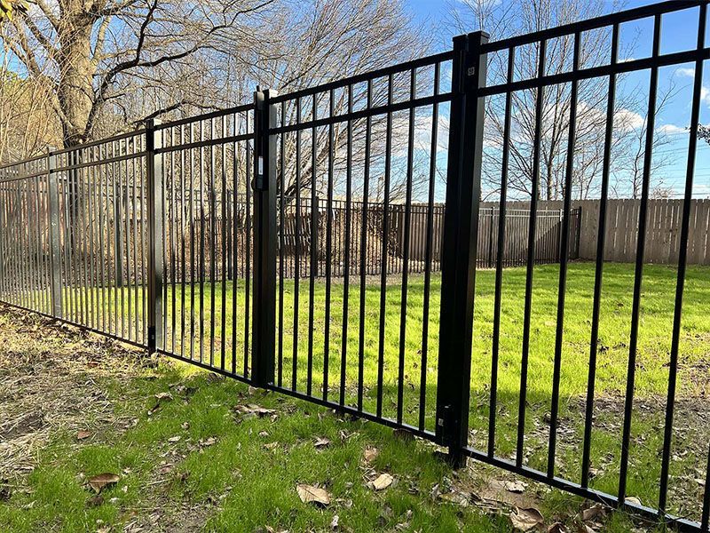 Black metal picket fence sections standing on a grassy lawn with trees in the background under a blue sky.
