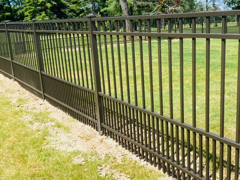 A brown metal fence with vertical slats and a puppy-picket design at the bottom, bordering a grassy yard.
