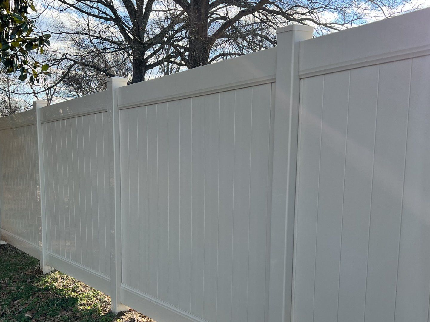 A modern white vinyl privacy fence stands outdoors with trees visible against a blue sky in the background.