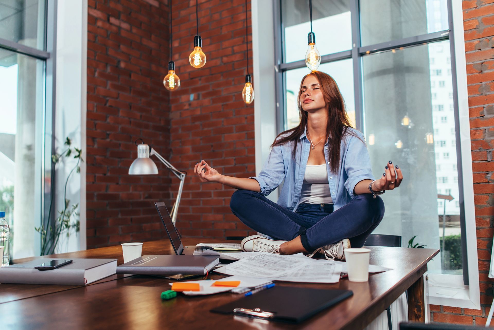 Mujer sentada con las piernas cruzadas sobre un escritorio, meditando con las manos en posición mudra.