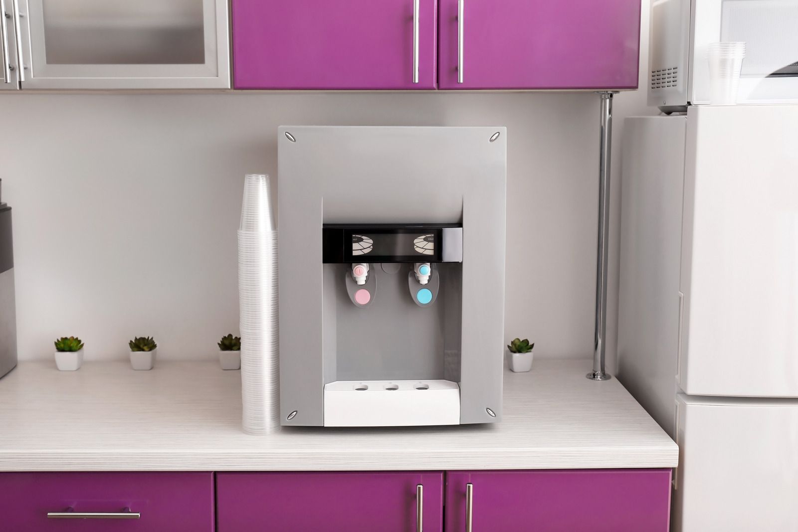 Countertop water dispenser in a kitchen with purple cabinets and a white refrigerator.